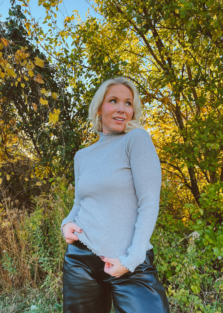 Woman in a gray top and black pants standing in a forest with autumn leaves.