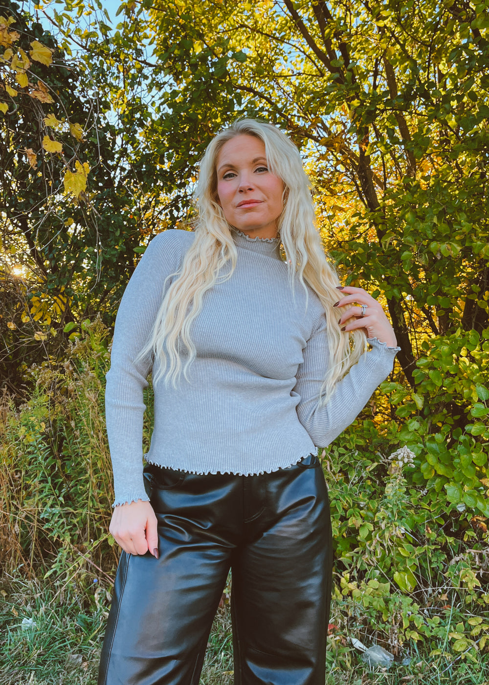 Woman in a gray long sleeve and black pants standing in a forest with trees and greenery in the background.