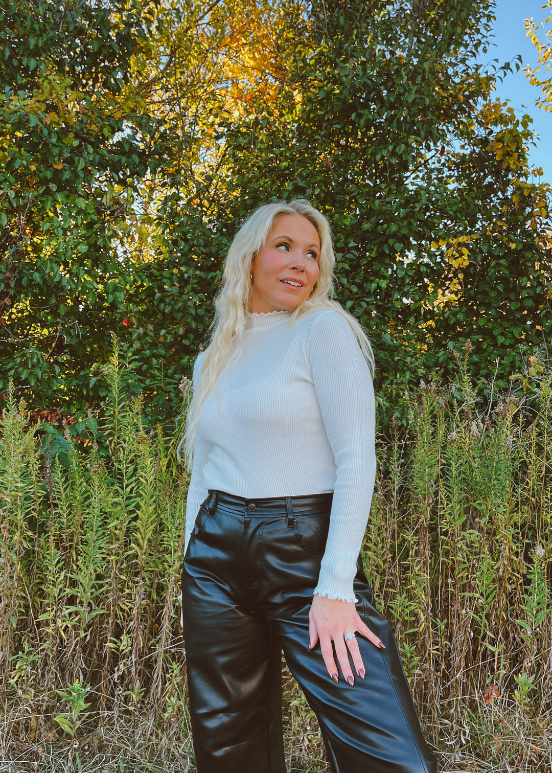 Woman in a white shirt and black pants standing in a natural setting with trees and plants.