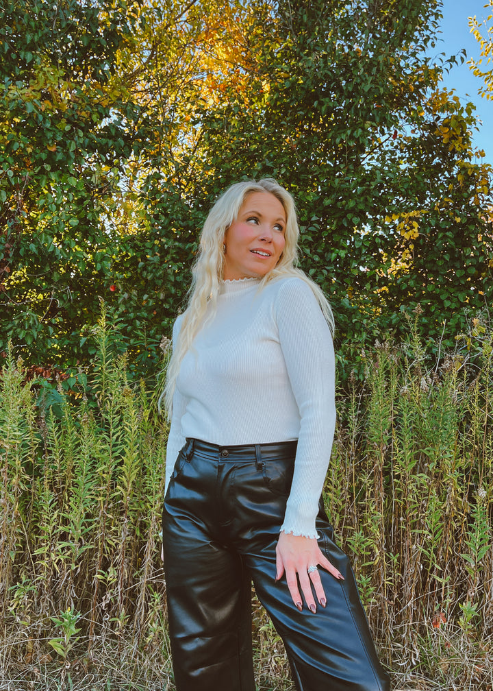 Woman in a white shirt and black pants standing in a natural setting with trees and plants.