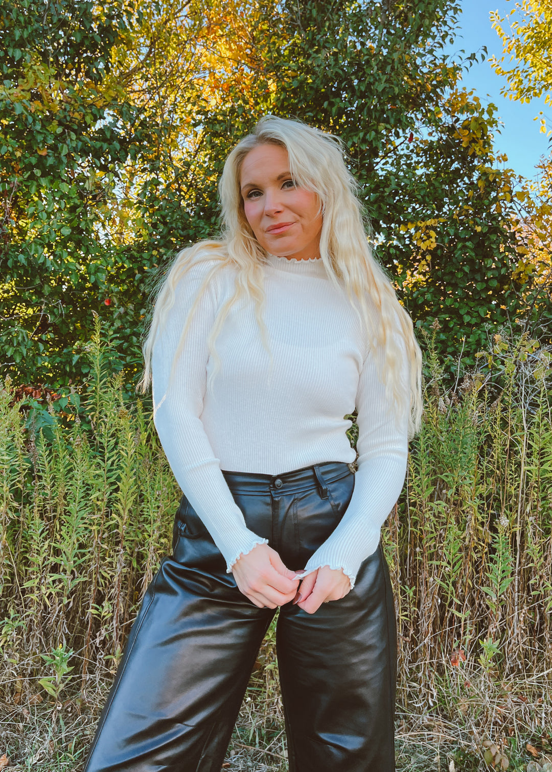 Woman wearing a white long-sleeve top and black leather pants standing in a natural setting with trees and grass.