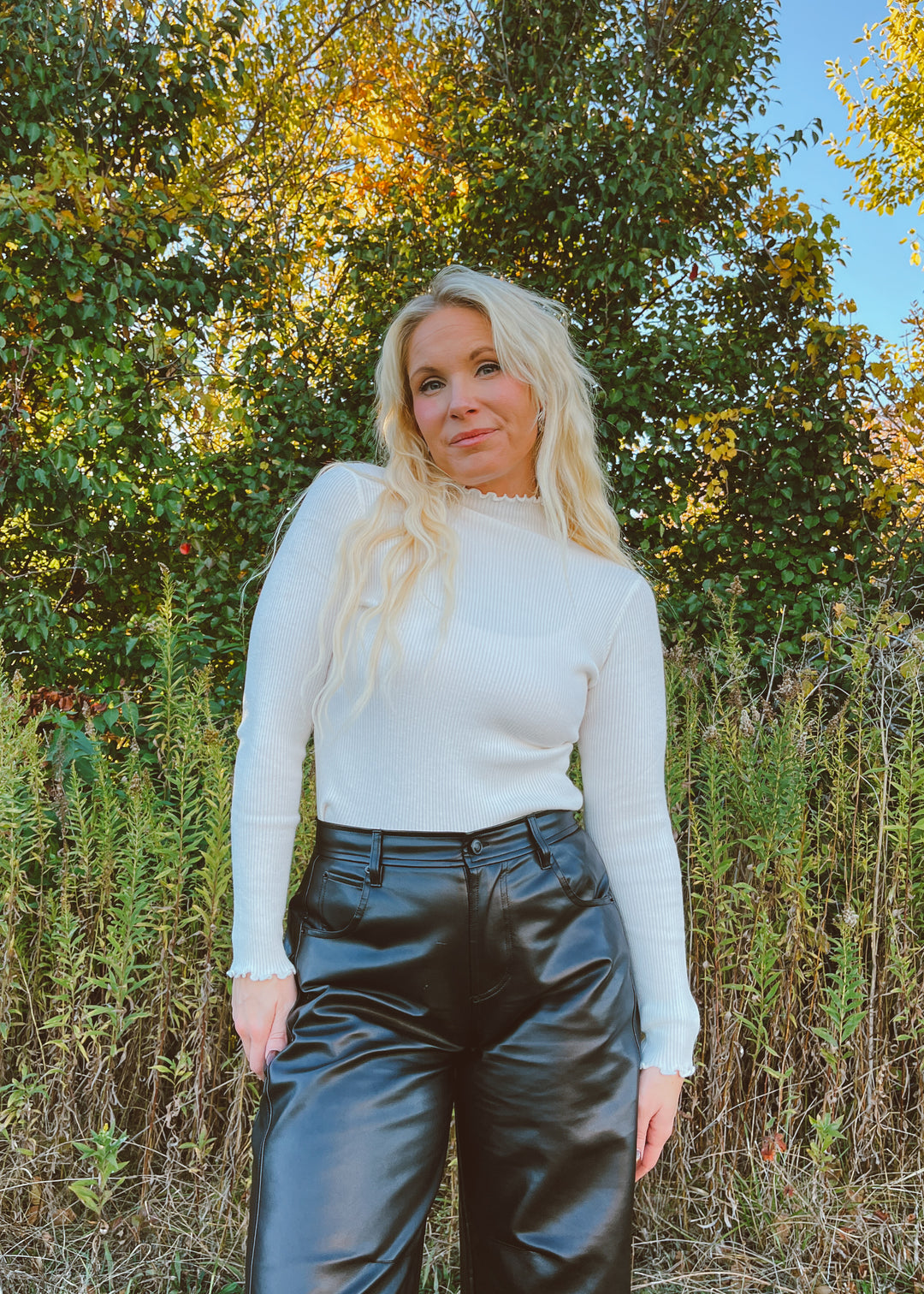Woman wearing a white long-sleeve top and black leather pants standing in a natural setting with trees and grass.