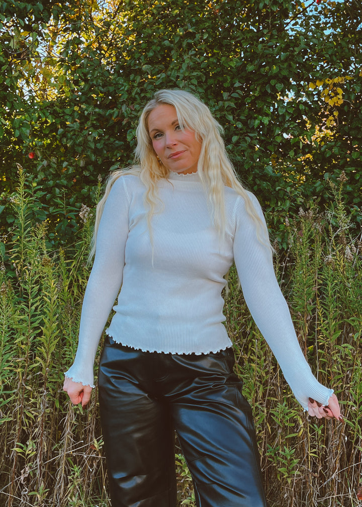 Woman wearing a white long-sleeve top and black pants standing in a natural setting with greenery.