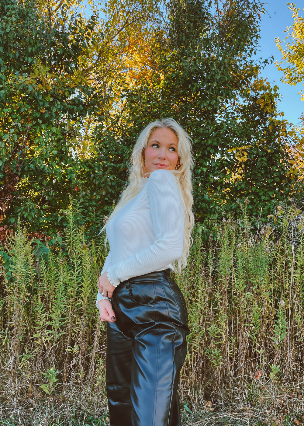 Woman in a white ribbed top standing outdoors with trees and foliage in the background