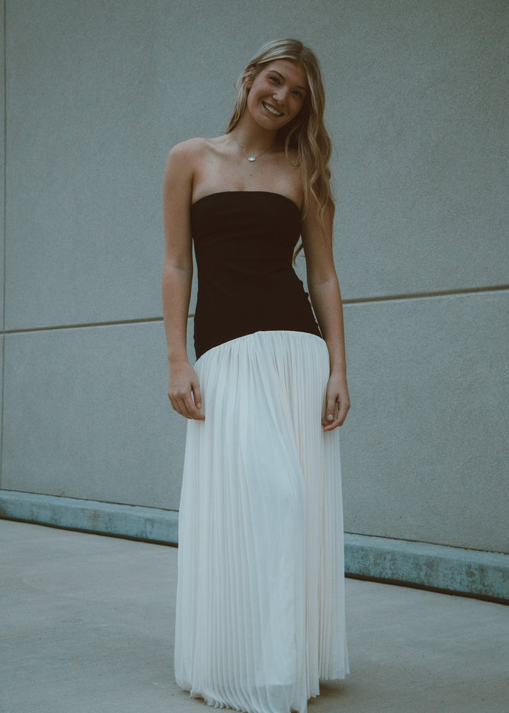 Woman wearing a black strapless and white pleated dress against a light gray wall.