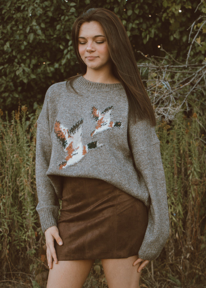 Woman wearing a gray sweater with a duck design and brown suede skirt outdoors