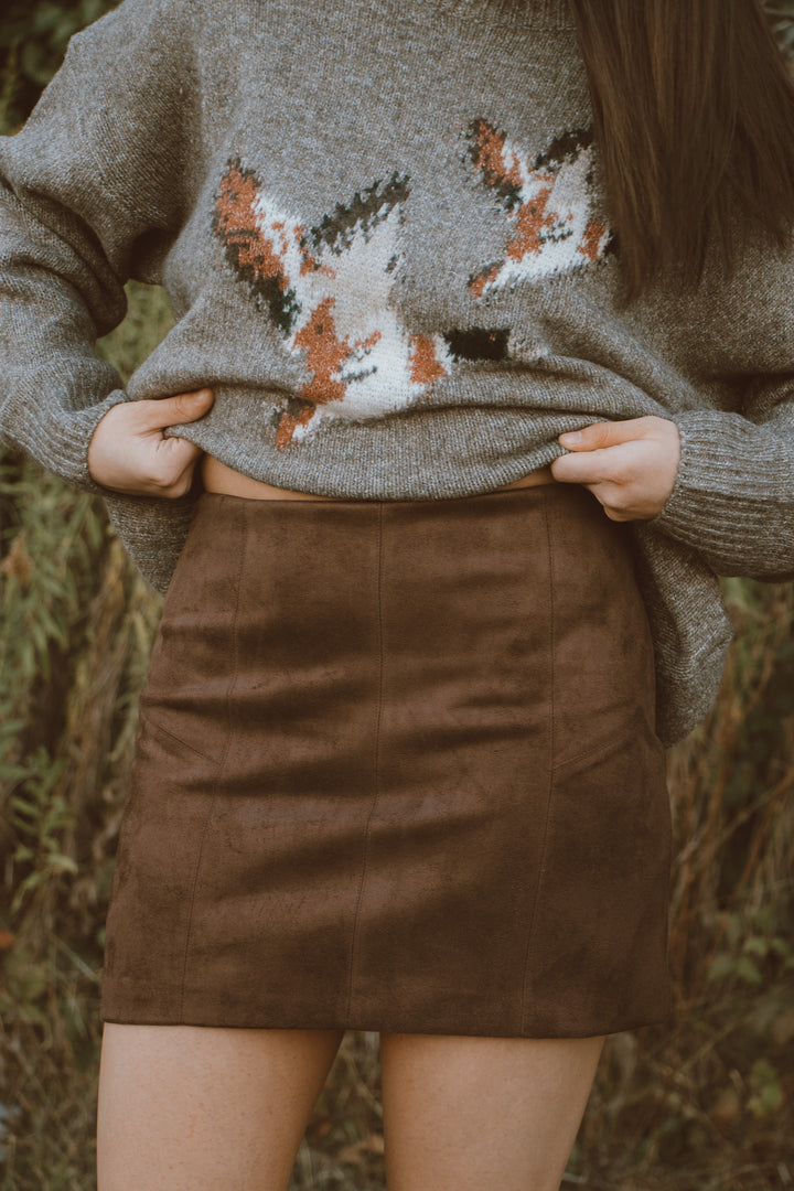 Person wearing a sweater with a duck design and a brown skirt outdoors.