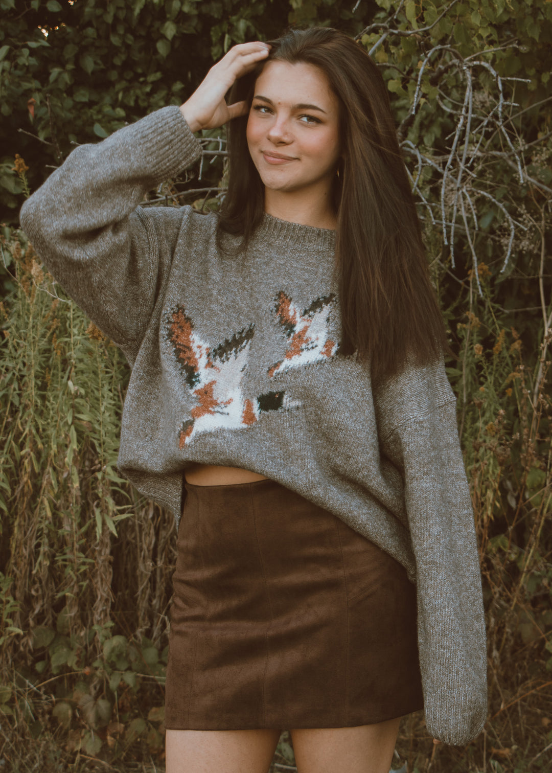 Woman wearing a gray sweater with duck patterns and a brown suede skirt outdoors.