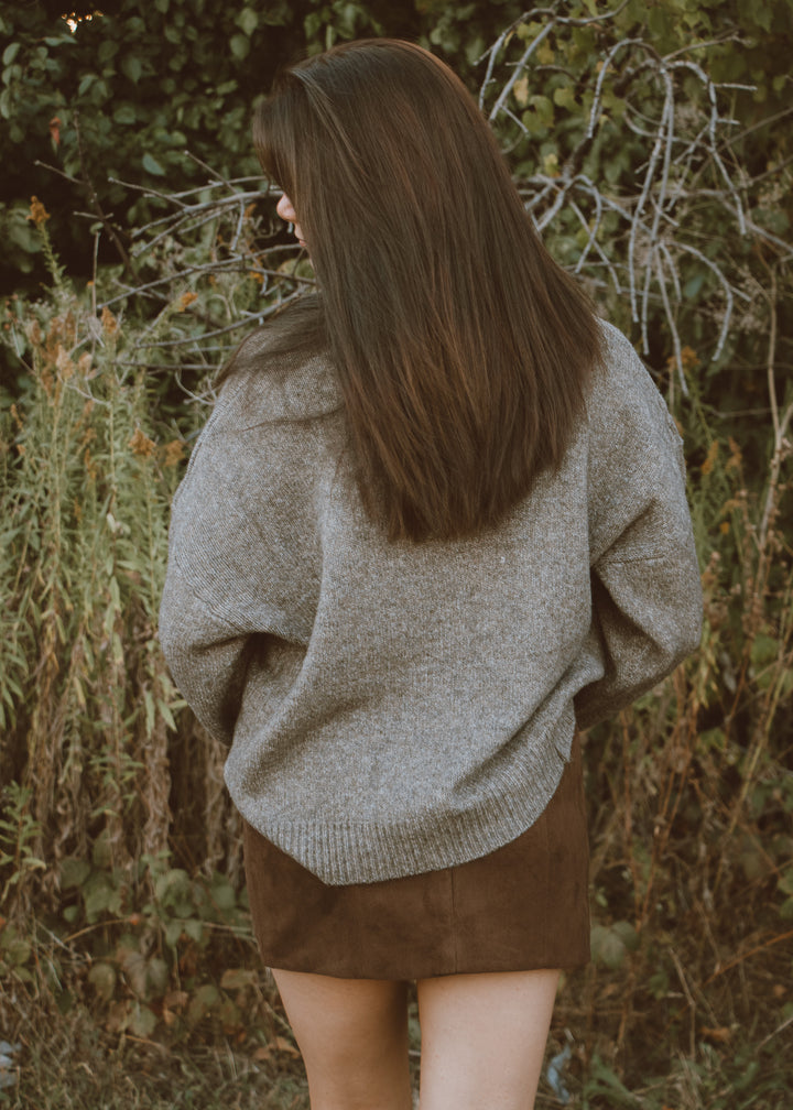 Person wearing a large gray sweater and brown skirt in a natural setting