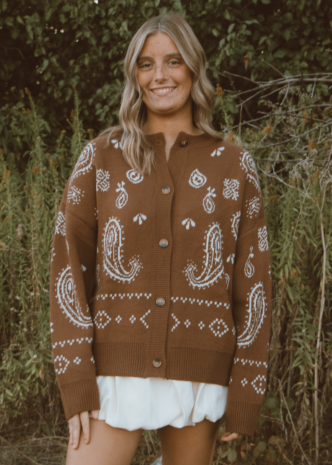 Woman wearing a brown cardigan with white patterns standing in a natural setting.