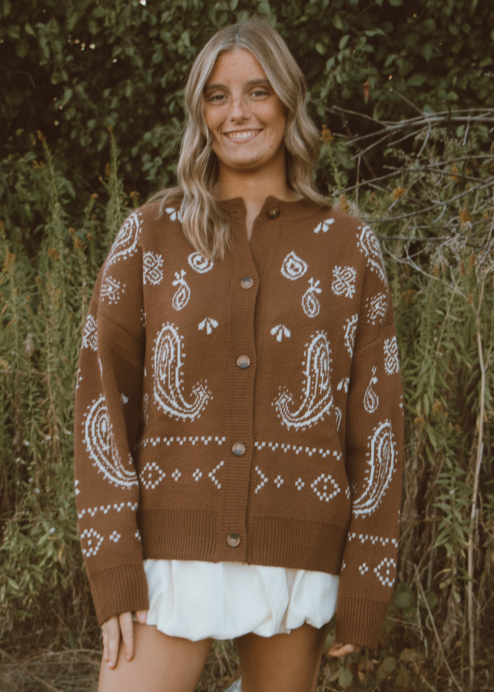 Woman wearing a brown cardigan with white patterns standing in a natural setting.
