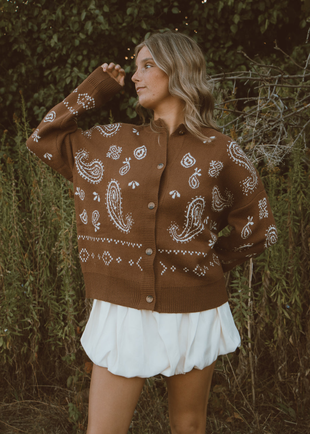 Woman wearing a brown patterned cardigan and a white skirt standing in a natural setting.
