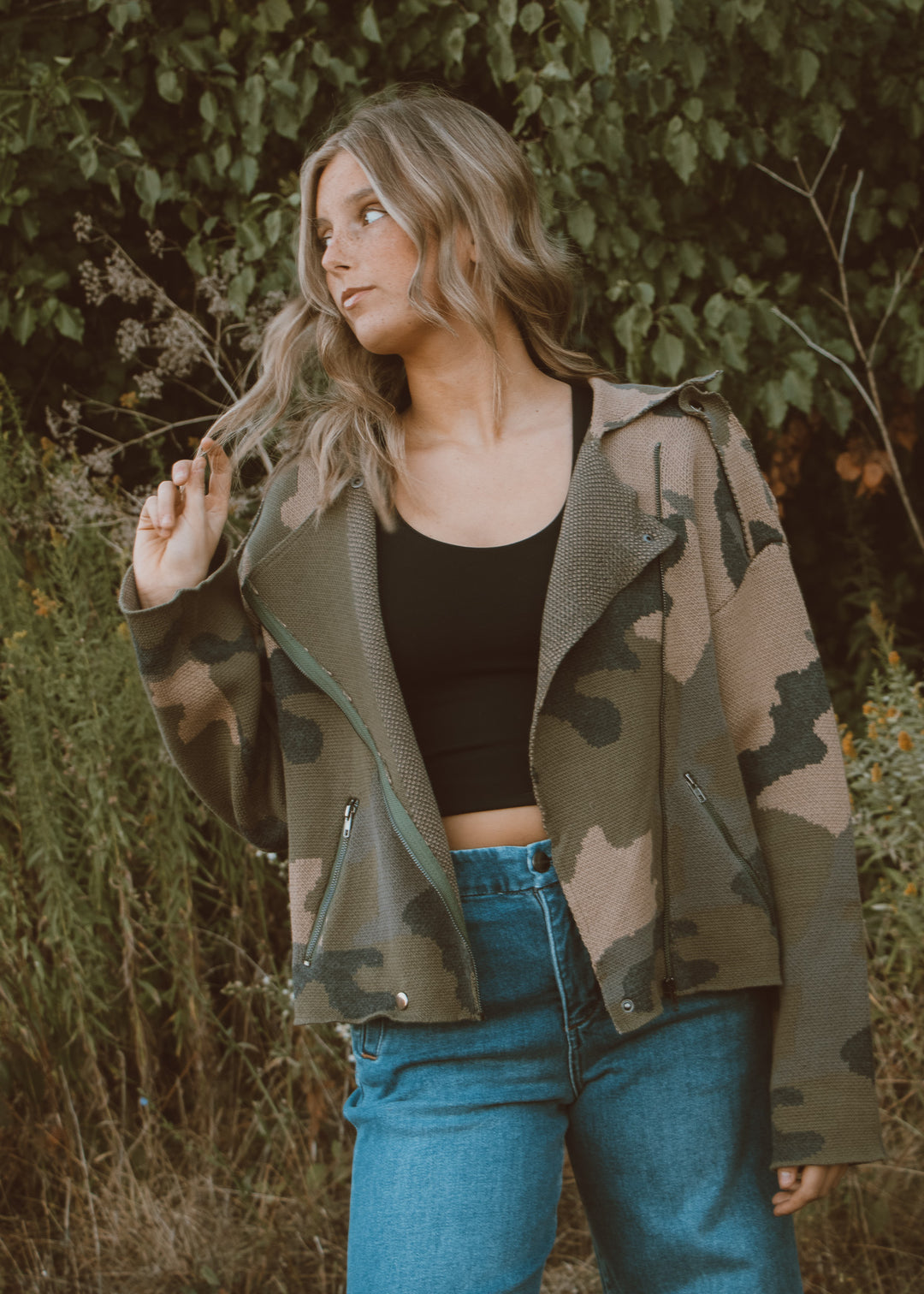 Woman wearing a camouflage jacket and blue jeans standing in a natural setting.