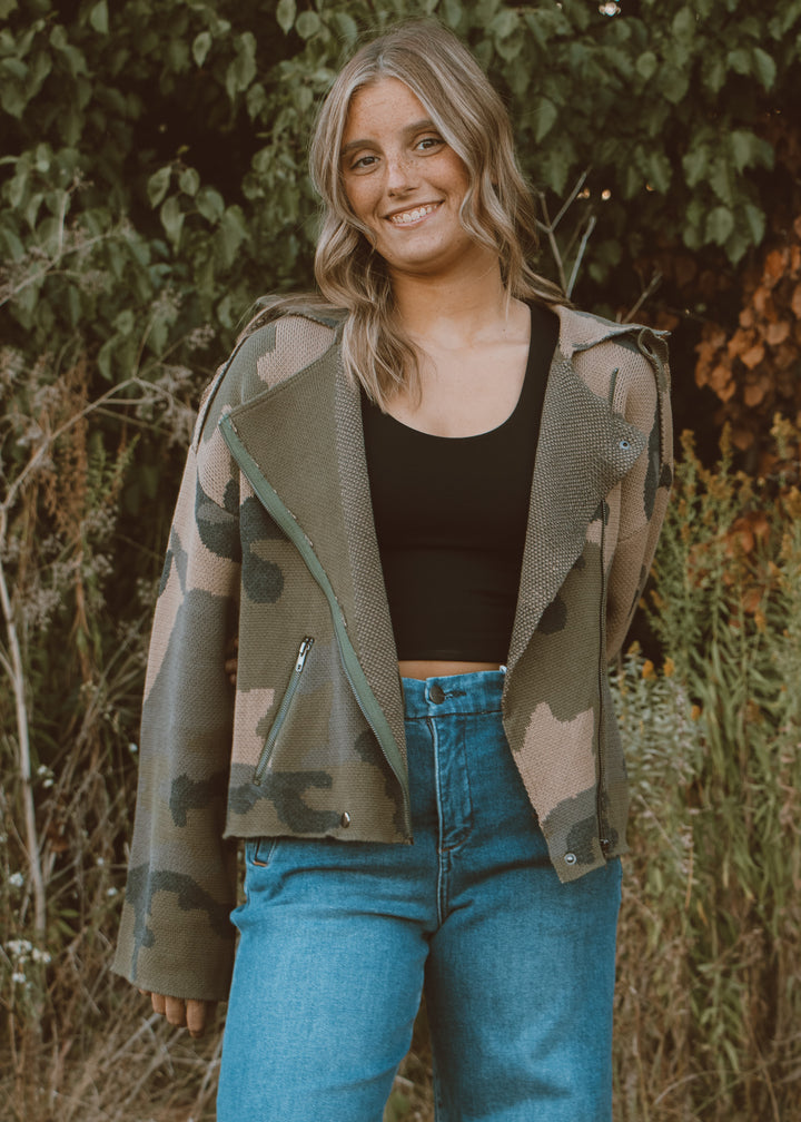 Woman wearing a camouflage jacket, black top, and blue jeans standing outdoors with greenery in the background