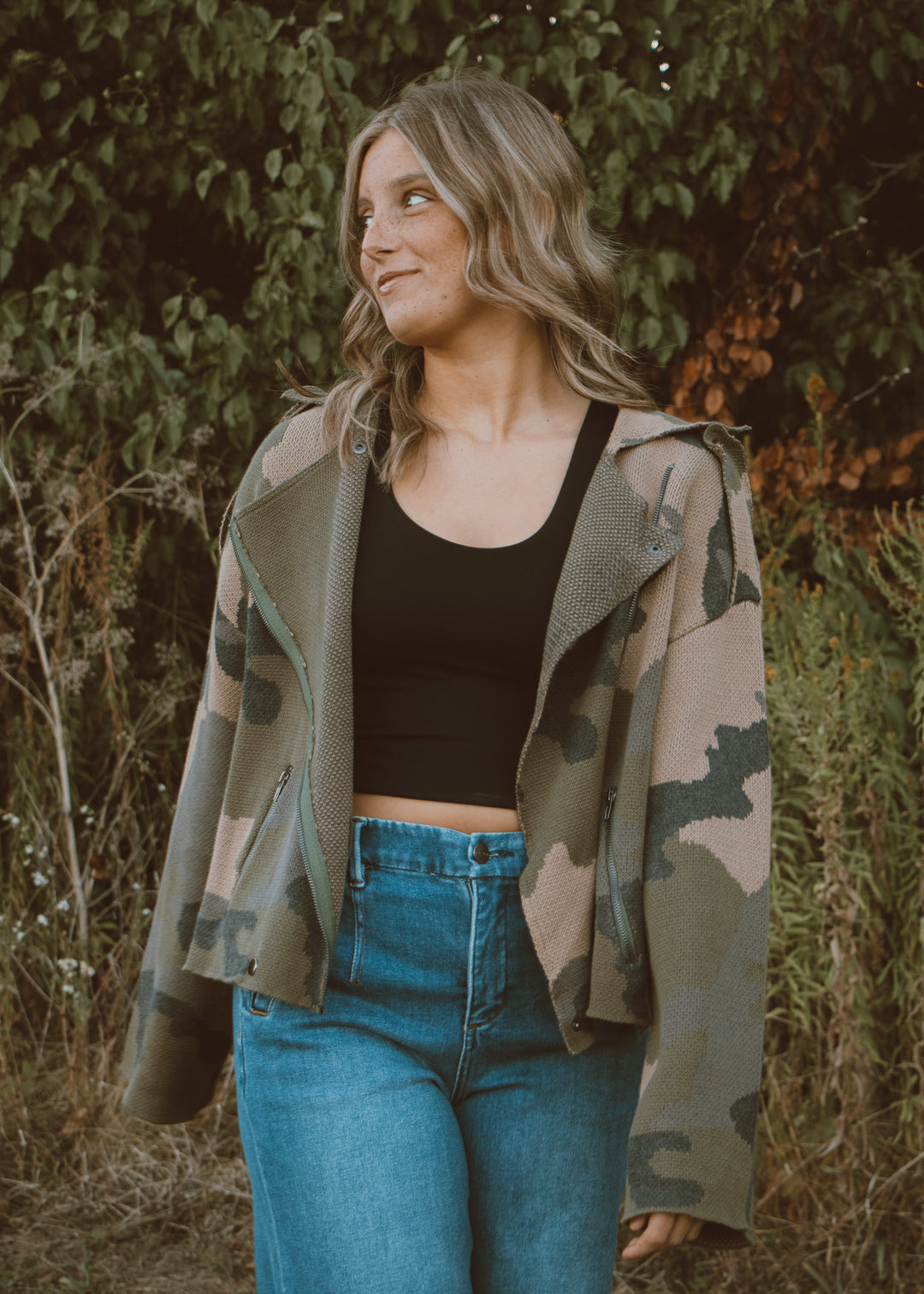 Woman wearing a camouflage jacket, black top, and blue jeans standing in a natural setting.