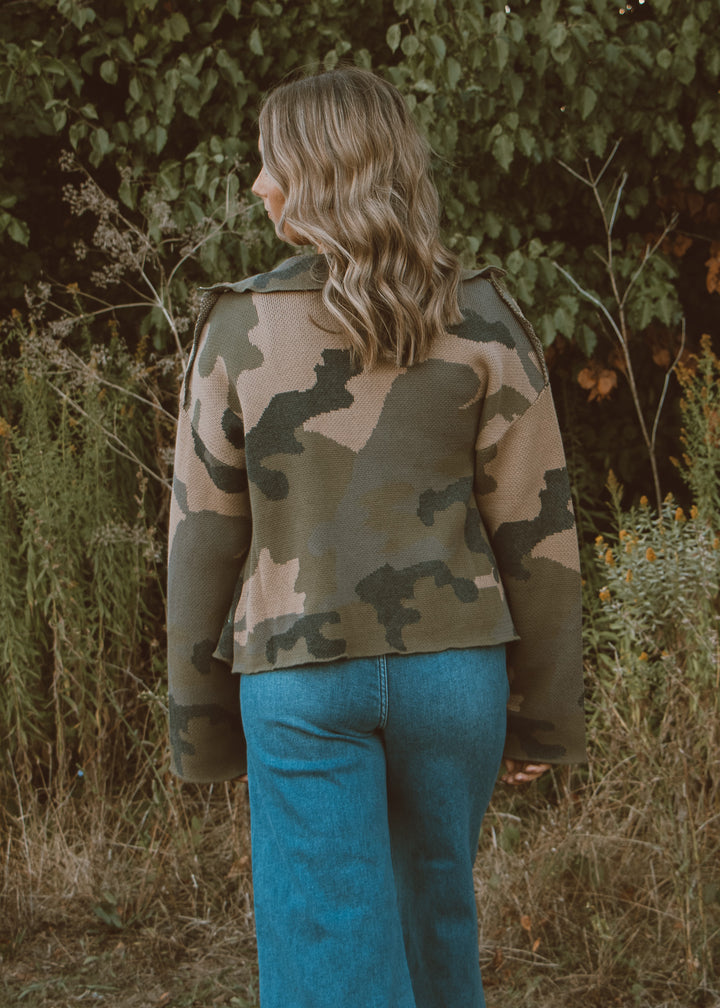 Person wearing a camouflage jacket and blue jeans standing in a natural setting with greenery.