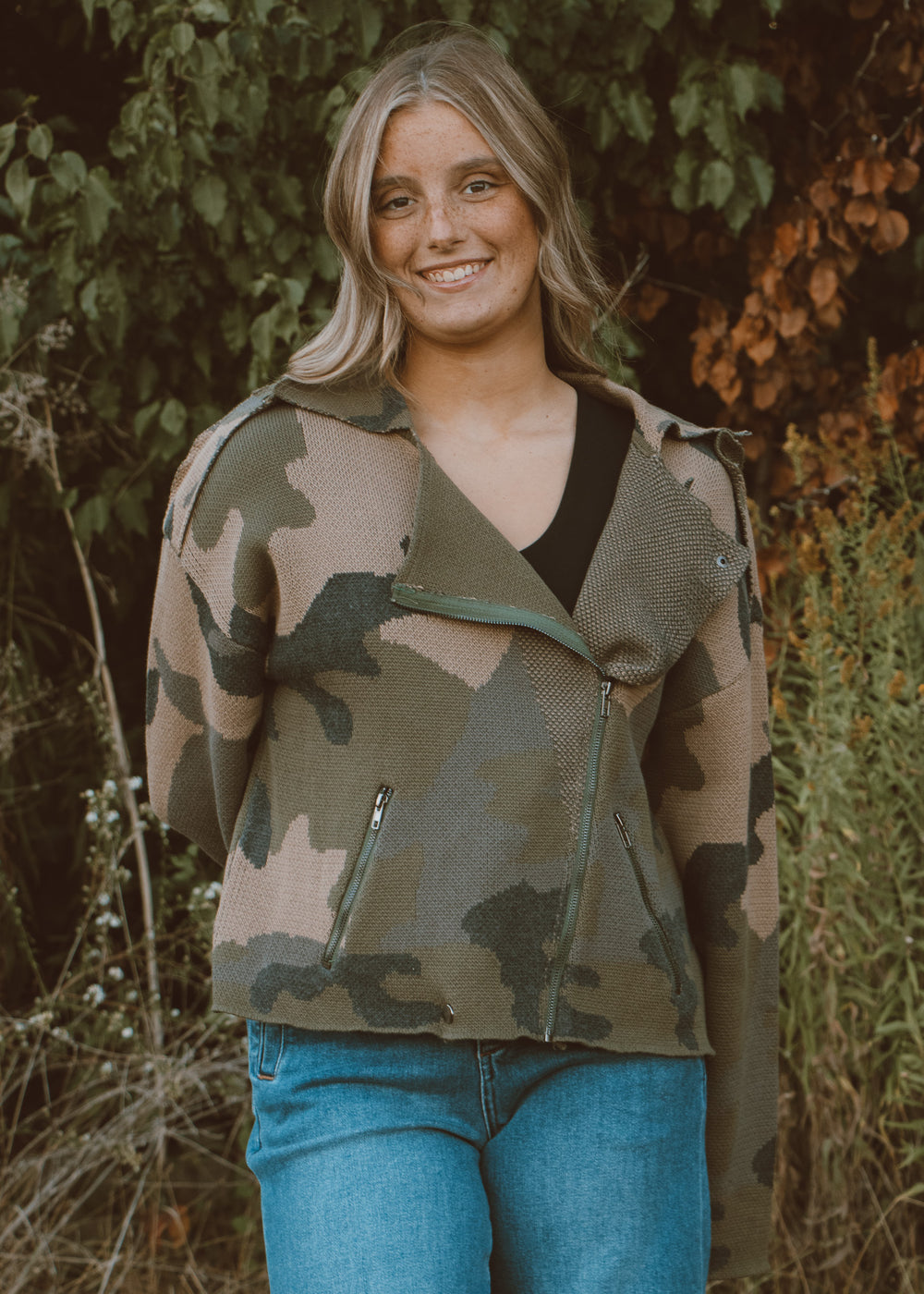 Woman wearing a camouflage jacket and blue jeans standing in a natural setting.
