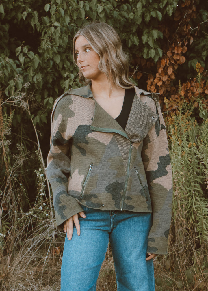 Woman wearing a camouflage jacket and blue jeans standing in a natural setting.