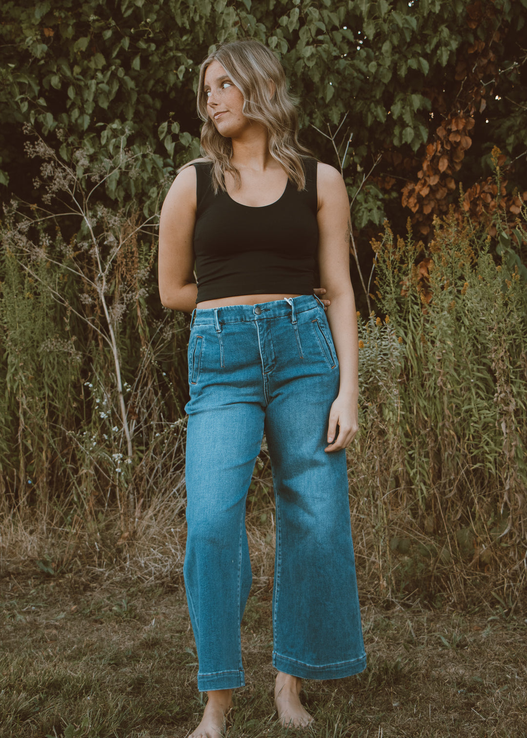 Woman wearing a black tank top and blue jeans standing in a natural setting with greenery.