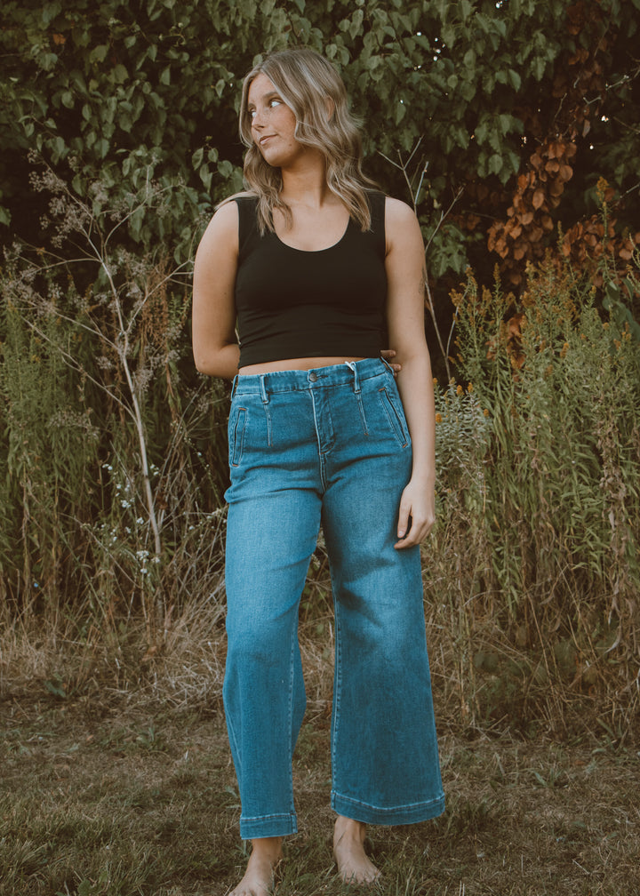 Woman wearing a black tank top and blue jeans standing in a natural setting with greenery.