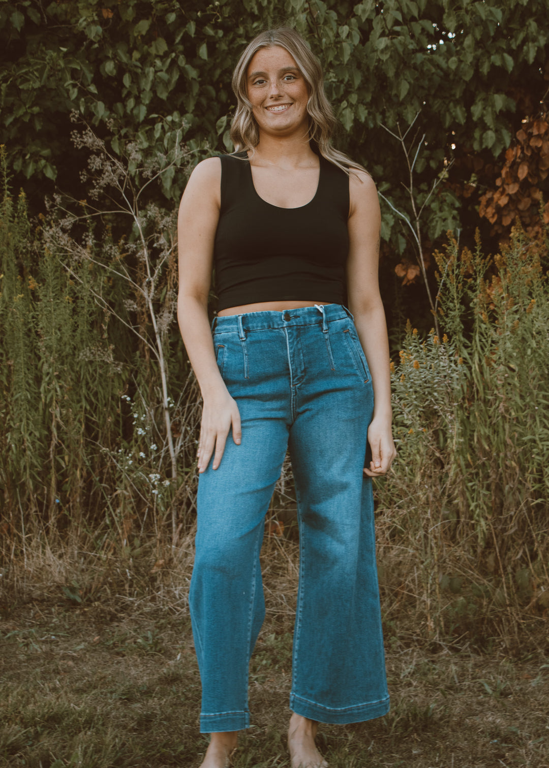 Woman wearing a black tank top and blue jeans standing in a natural setting with greenery.