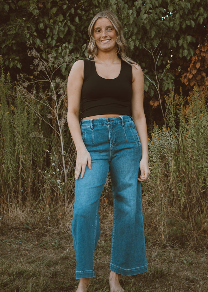 Woman wearing a black tank top and blue jeans standing in a natural setting with greenery.