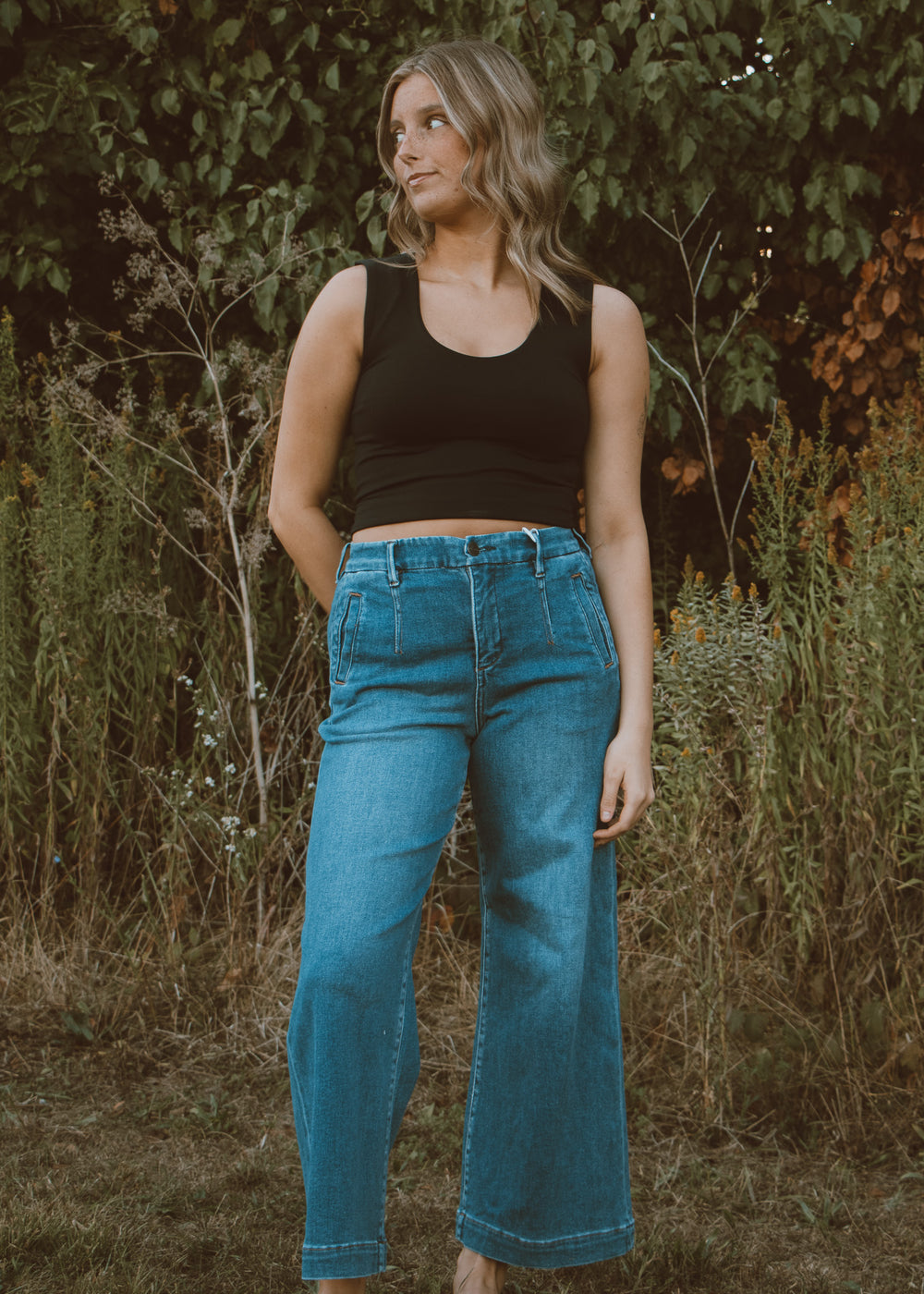 Woman wearing a black tank top and blue jeans standing in a natural setting with greenery.