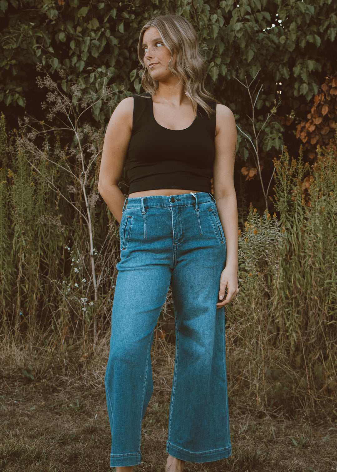 Woman wearing a black tank top and blue jeans standing in a natural setting with greenery.