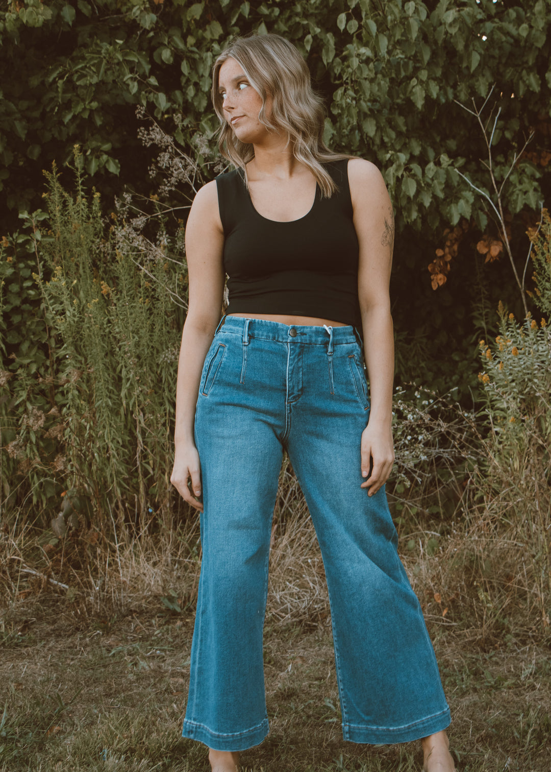 Woman wearing a black tank top and blue jeans standing in a natural setting with greenery.