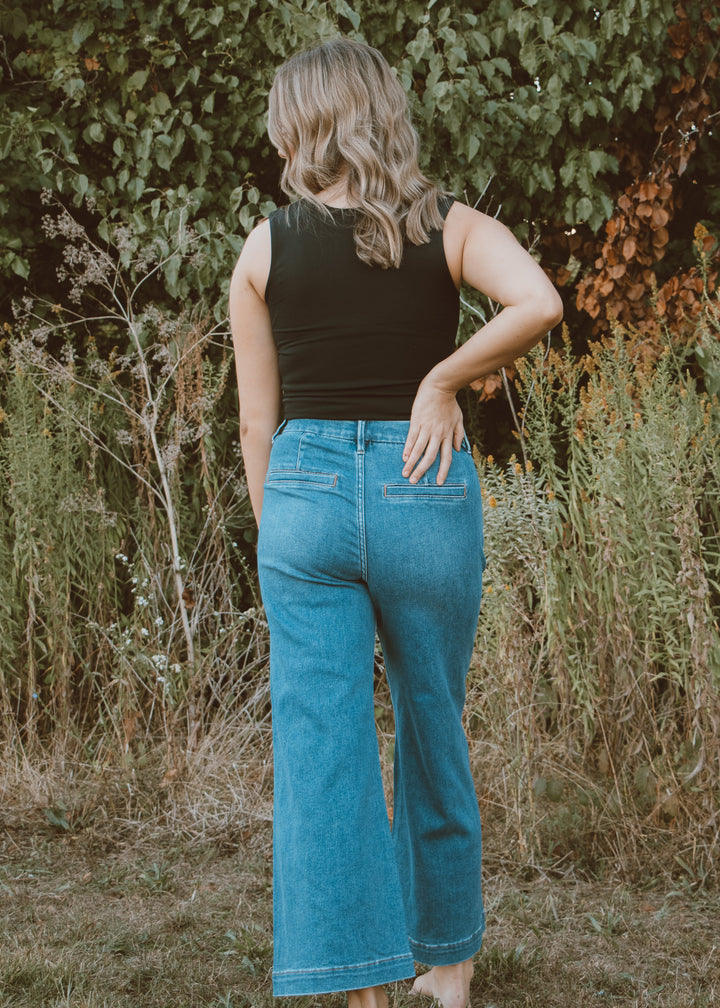 Person wearing a black tank top and blue jeans standing in a natural setting with greenery.