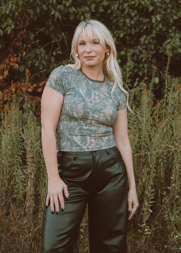 Woman wearing a camo top and dark pants standing in front of greenery