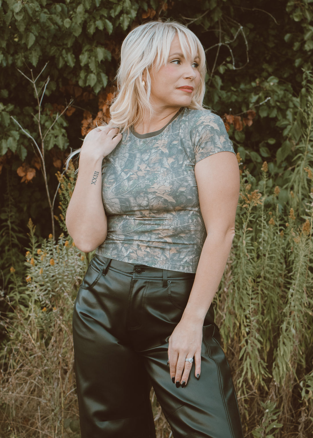 Woman wearing a camo top and black leather pants standing in a natural setting