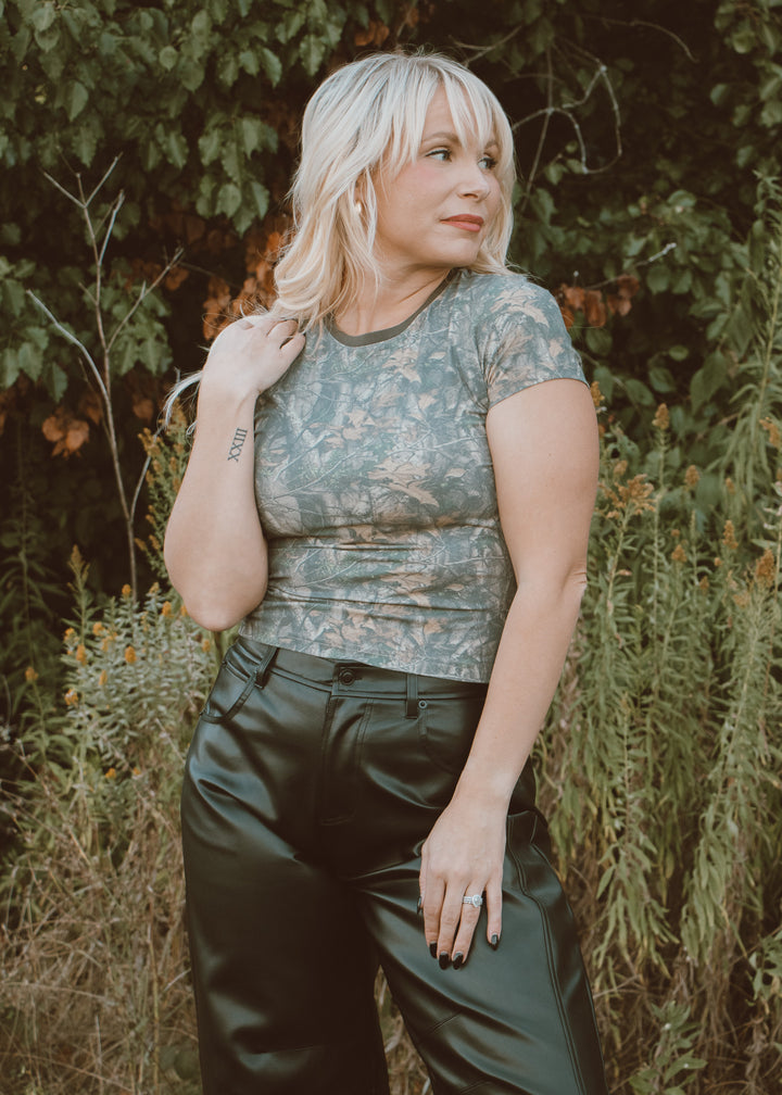Woman wearing a camo top and black leather pants standing in a natural setting