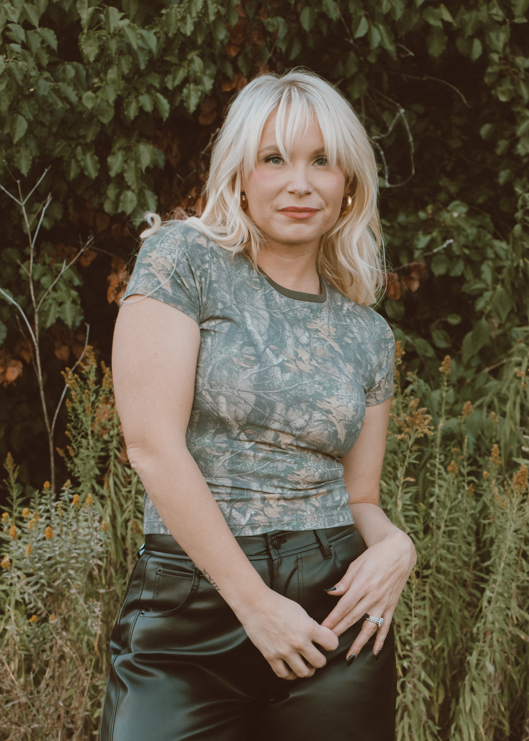 Woman wearing a camo shirt and black pants standing in a natural setting with greenery.