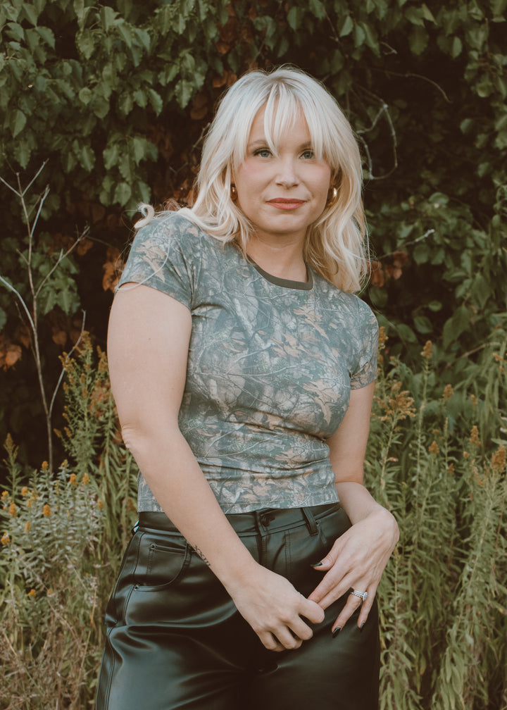 Woman wearing a camo shirt and black pants standing in a natural setting with greenery.