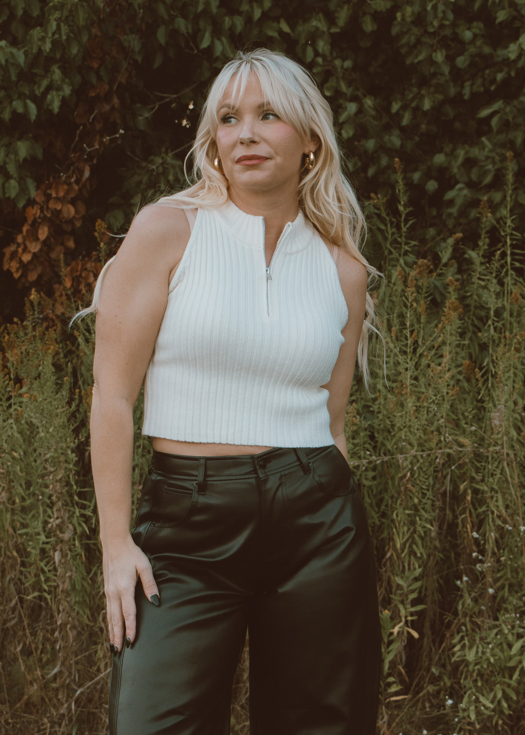 Woman wearing a white sleeveless top and dark pants standing in front of green foliage.
