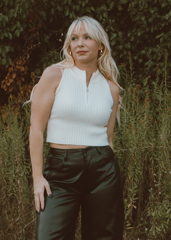 Woman wearing a white sleeveless top and dark pants standing in front of green foliage.