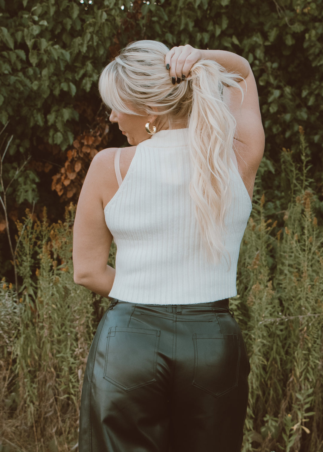 Woman with blonde hair tied back, wearing a white top and black leather pants, standing in a natural setting.
