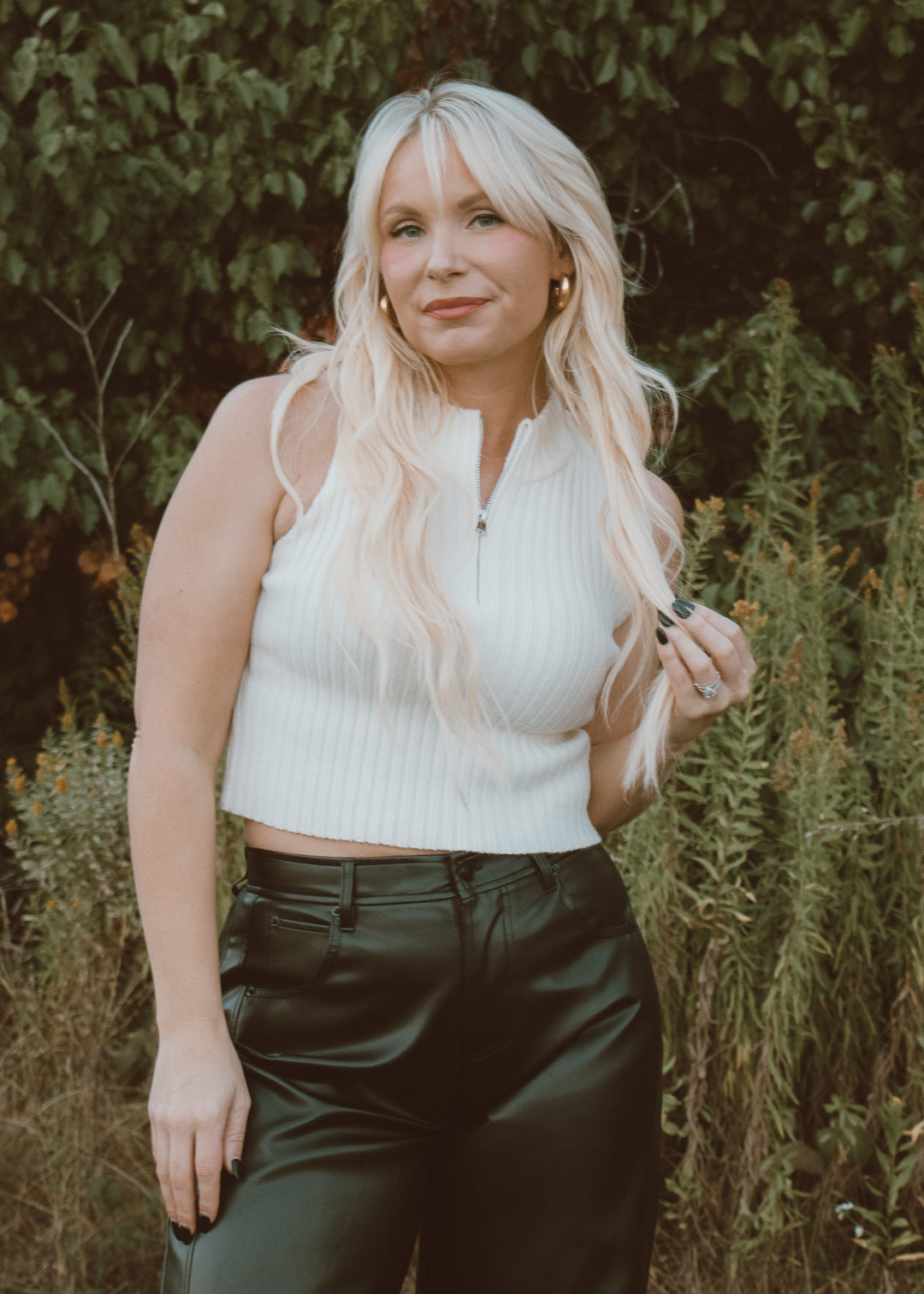 Woman with long blonde hair wearing a white sleeveless top and dark pants outdoors.