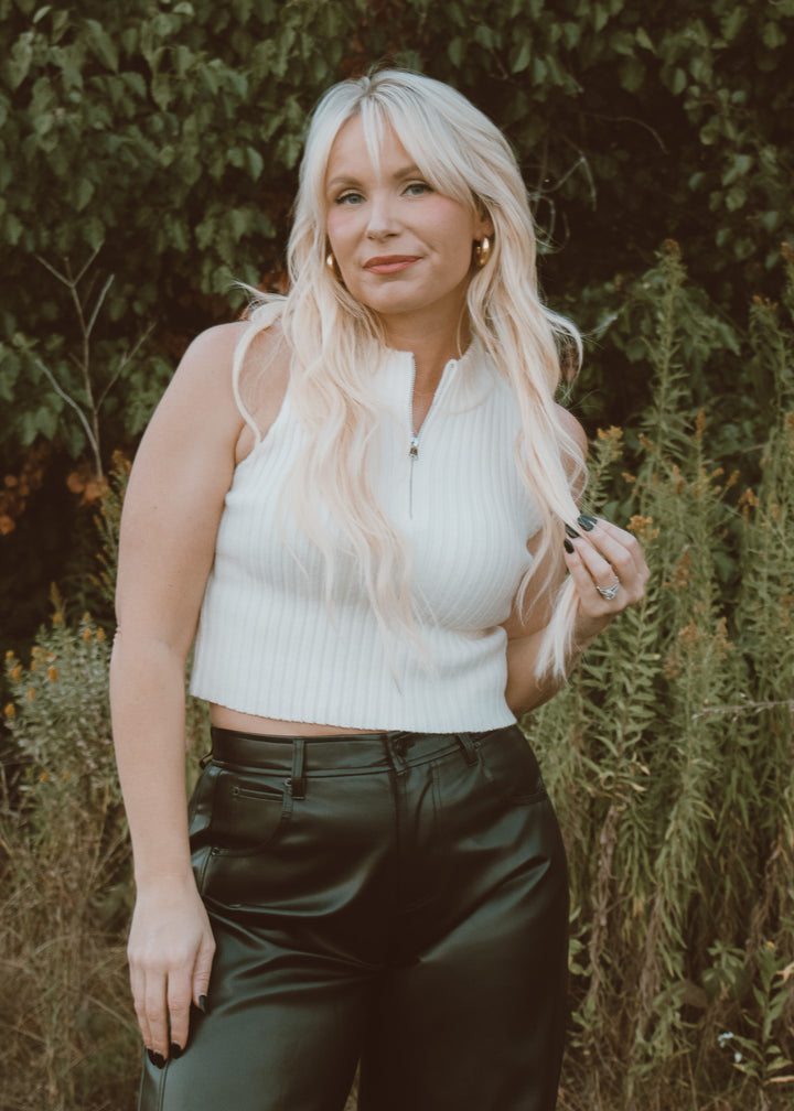 Woman with long blonde hair wearing a white sleeveless top and dark pants outdoors.