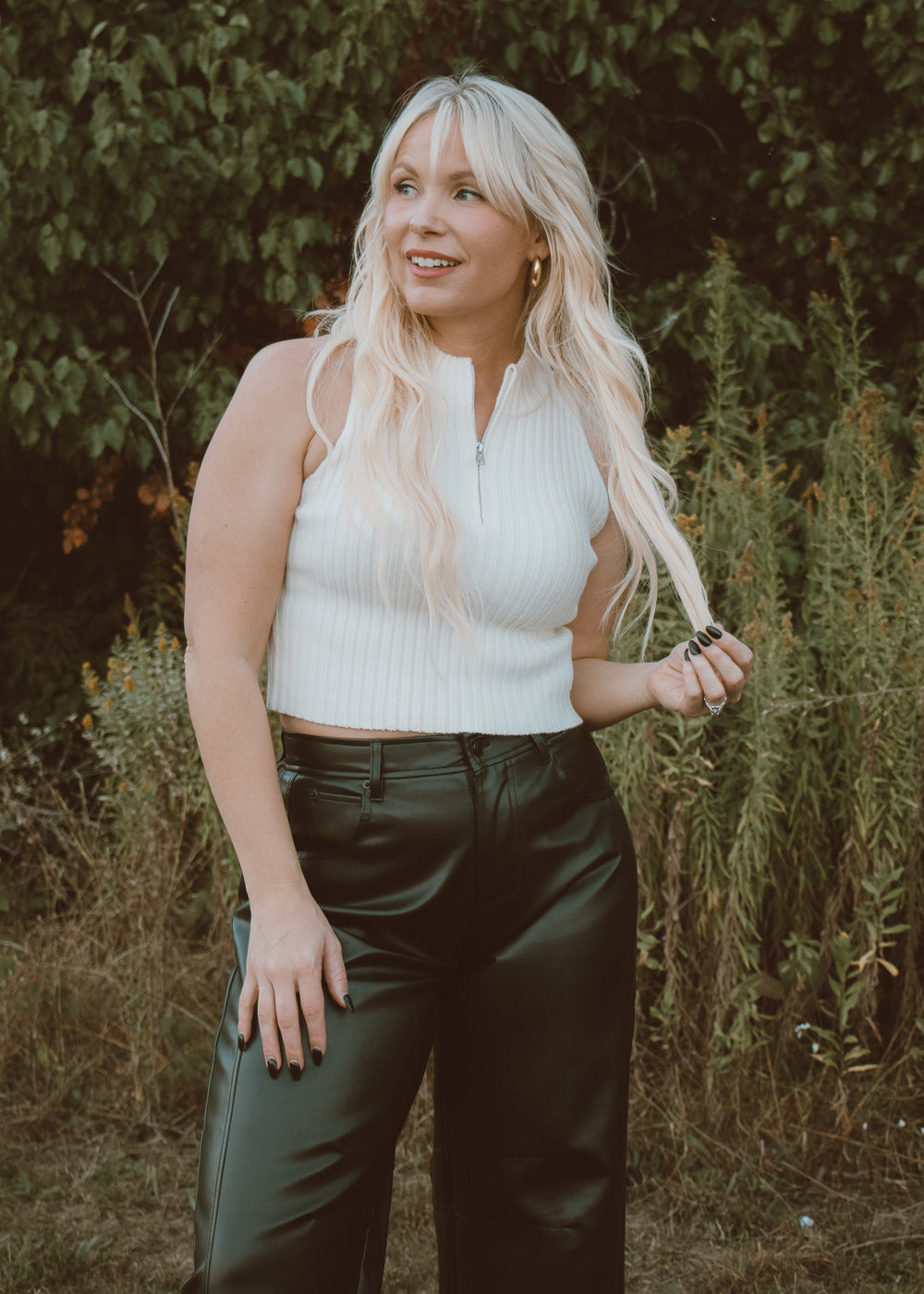 Woman with long blonde hair wearing a white sleeveless top and black leather pants standing in a natural setting.