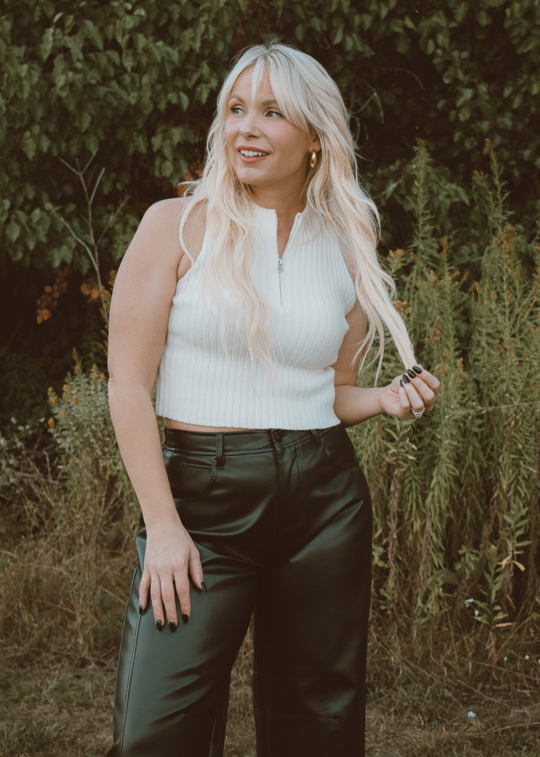 Woman with long blonde hair wearing a white sleeveless top and black leather pants standing in a natural setting.