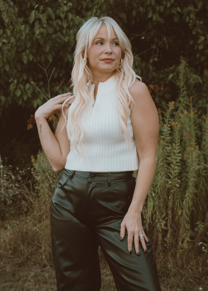Woman wearing a white sleeveless top and dark pants standing in a natural setting.