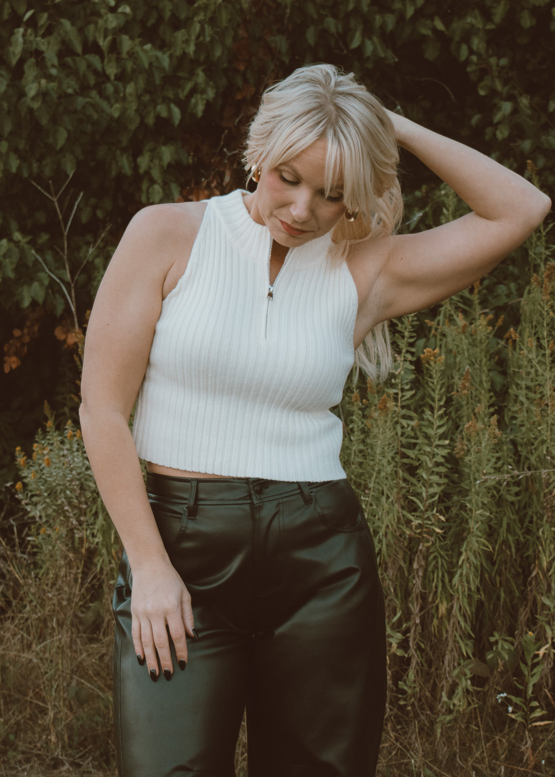 Woman wearing a white sleeveless top and dark pants standing in a natural setting.