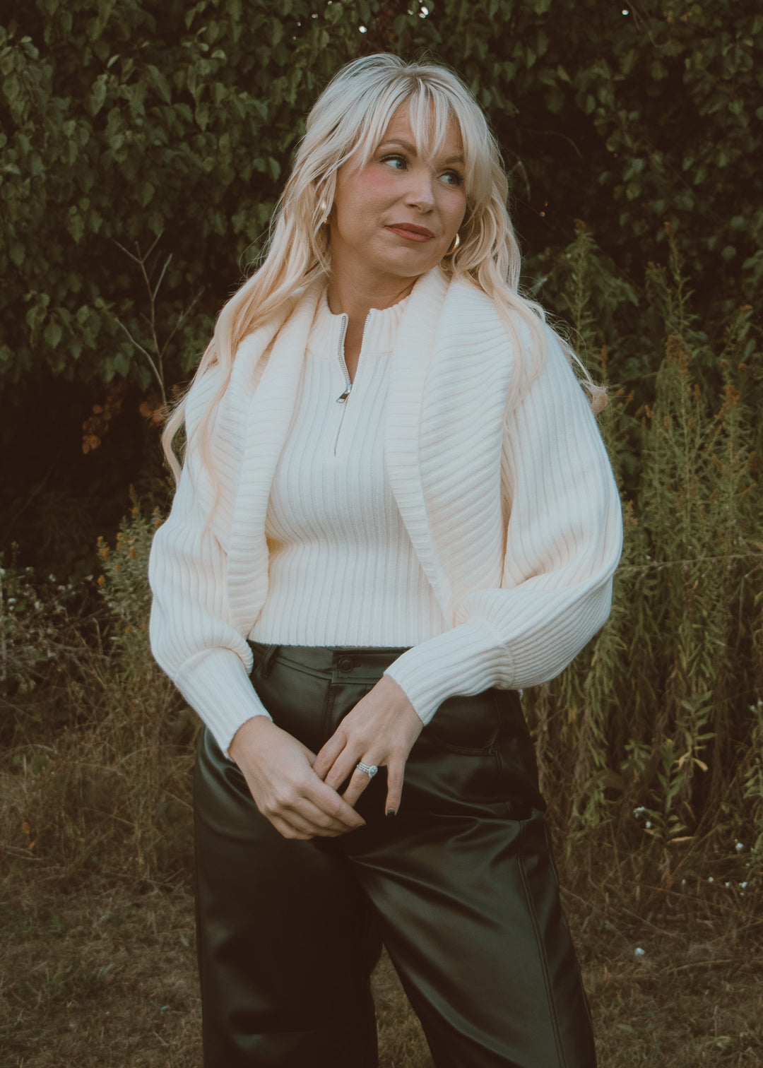 Woman wearing a white sweater and dark pants standing in a natural setting.