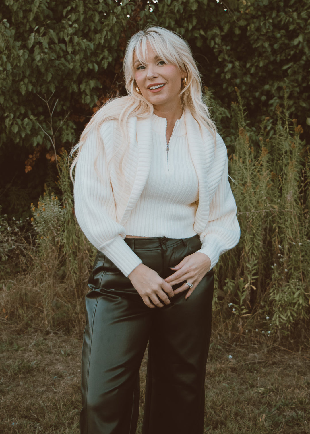 Woman wearing a white sweater and black leather pants standing outdoors with greenery in the background