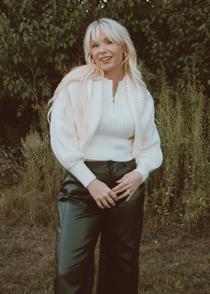 Woman wearing a white sweater and black leather pants standing outdoors with greenery in the background
