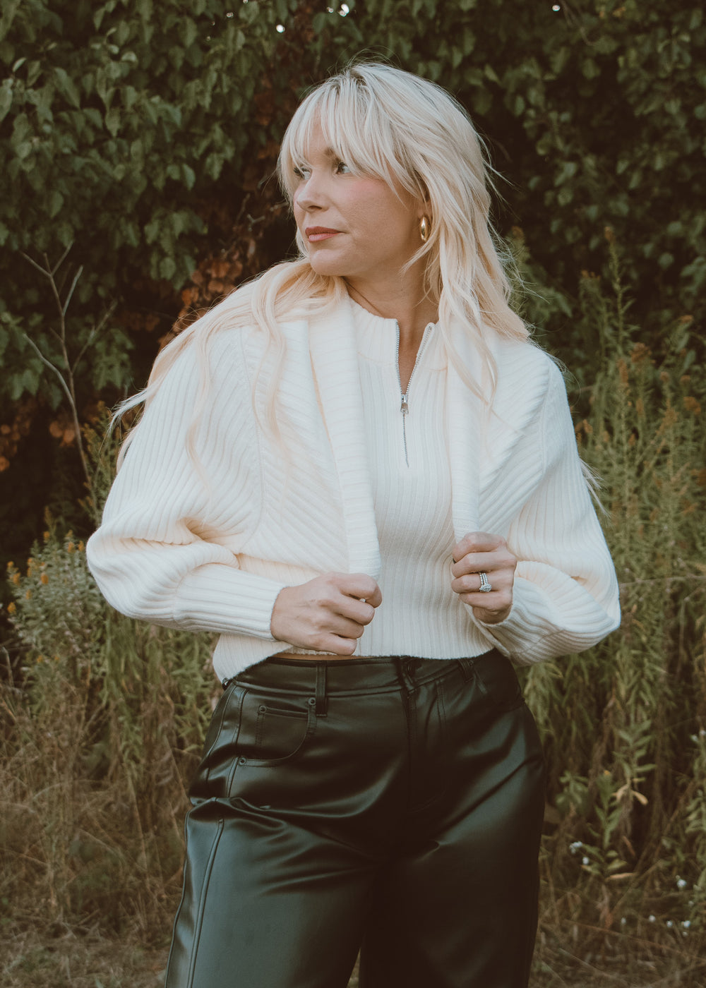 Woman wearing a white shrug sweater over a white shirt with dark pants in a natural setting.