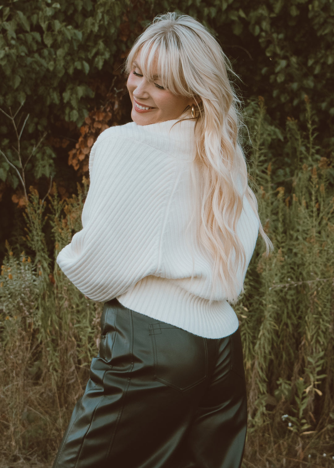 Woman wearing a white sweater and dark pants standing in a natural setting with greenery.