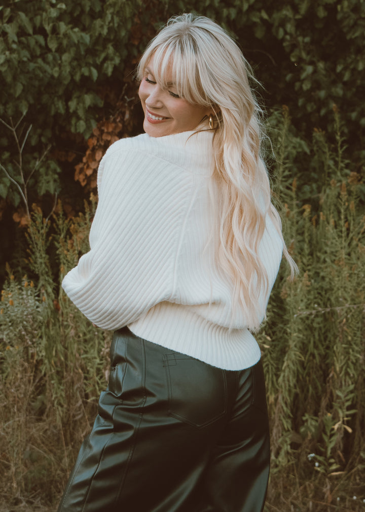 Woman wearing a white sweater and dark pants standing in a natural setting with greenery.